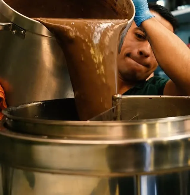 A cook pouring the thick hot chocolate from one pot into another.